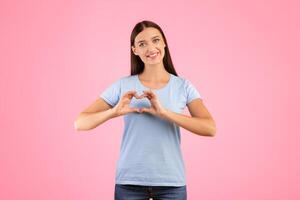 Love And Affection. Portrait of cute woman making heart shape with her hands and fingers near chest, smiling to camera, posing over yellow background in studio, copy space. Romantic Concept. photo