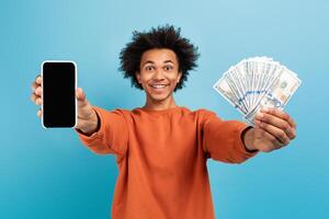 A young African American man smiles widely while holding a smartphone in one hand and a bundle of cash in the other against a bright blue background, reflecting joy and accomplishment. photo