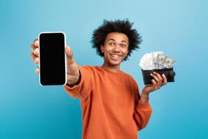 A smiling African American man holds a smartphone in one hand and a wallet filled with cash in the other. He exudes confidence and excitement in a vibrant blue setting. photo