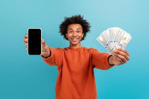 A young African American man with curly hair smiles widely while holding a smartphone in one hand and a stack of cash in the other, embodying a cheerful lifestyle and achievement. photo