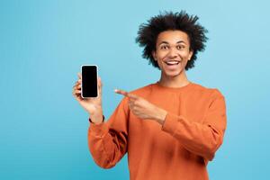 In a bright blue setting, a joyful African American man points at a smartphone with a big smile. His playful expression and trendy outfit highlight a modern lifestyle and tech engagement. photo