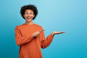 A cheerful African American man in an orange sweater shows enthusiasm as he points at his empty hand with a smile, copy space photo