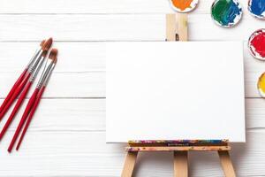 Artist workspace featuring red paintbrushes, various paint palettes, and a blank canvas on an easel ready for creative expression photo