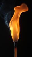 A vibrant flame rises and curls elegantly, surrounded by wisps of smoke in a striking contrast against darkness photo