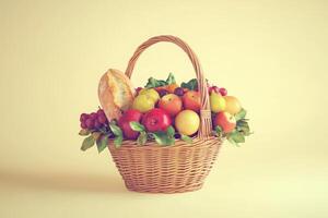 A rustic wicker basket filled with a colorful assortment of fruits including apples, oranges, and grapes, alongside a crusty loaf of bread. The backdrop is a soft yellow, enhancing the vibrant colors. photo