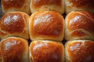 Soft, golden-brown rolls cool on a wire rack in a kitchen. The warm lighting highlights their shiny surfaces, making them look inviting and freshly baked. Perfect for a meal or snack. photo