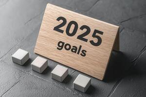 A wooden sign displays the year 2025 along with the word goals. Next to it are four gray blocks, symbolizing the steps or milestones towards achieving these goals, on a textured surface. photo