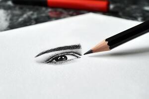 A pencil is poised above a realistic sketch of an eye on white paper, showcasing intricate details. Red art supplies are visible, suggesting an artistic workspace during the day. photo