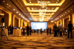 A spacious banquet hall is softly lit with golden tones, featuring elegantly dressed guests mingling. The atmosphere is festive as a large screen displays visuals, creating an inviting ambiance. photo