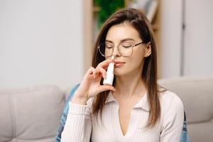 Young woman wearing casual clothing, using a nasal spray to facilitate breathing, sitting on a sofa in a home interior photo