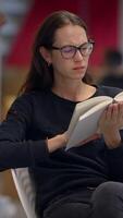 un mujer es sentado cómodamente en un moderno biblioteca ajuste mientras profundamente leyendo un libro video