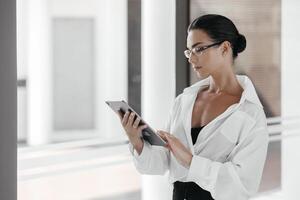 Attractive businesswoman using a digital tablet while standing in front of windows in an office building. Free copy space, banner, ads. female surf web read news or chat on tablet device, technology. photo