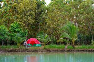 Senior man sits in front of a tent.Red camping tent in the riverside forest at night photo