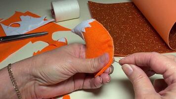 Woman sews up felt toy filled with padding polyester at her workplace for hobby at home. Making handmade felt toys for small children. Creating fox toy from orange felt with background of details video