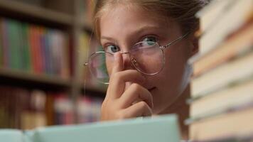 Close up clever smart intelligent Caucasian little girl playful schoolgirl child school kid pupil learner student in glasses eyeglass looking at camera reading book literature inside library education video
