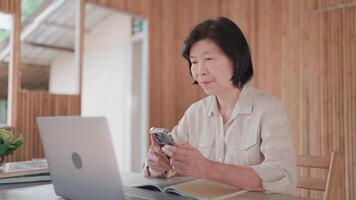 Senior Asian woman using smartphone and laptop in bamboo house to learn and research online. Self development, digital connectivity, gadgets, online learning concept. video