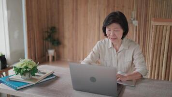 Senior woman is using pen to take notes and using laptop to talk on call and there is a vase of flowers and books on the table in bamboo house. Online learning. video