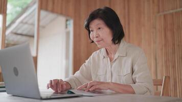 Senior woman having call using laptop and reading book at table in bamboo house. Online learning. call with family. Searching information on internet. Senior citizen learning online. video
