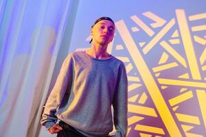 A young man in a gray sweater stands in front of a colorful background, looking directly at the camera photo