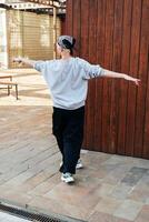 A person wearing a grey sweatshirt and black pants walks away from a wooden wall with their arms outstretched photo