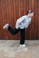A man in a grey sweater and black pants is doing a dance move in front of a wooden wall photo