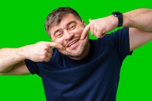 A smiling man playfully poking his cheeks against a vibrant green background while showing off his teeth photo