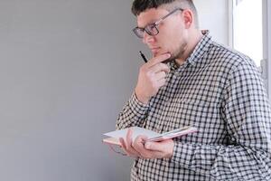 man in plaid shirt sitting at windowsill , thinking and writing in a notebook photo