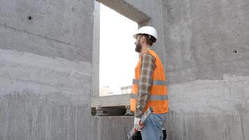 Builder repairman, foreman in protective helmet and vest stands at workplace in building and holds ruler and toolbox in hands video