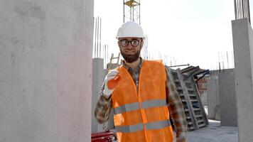 Builder repairman, foreman in protective helmet and vest stands at his workplace in the building under construction and shows ok gesture video
