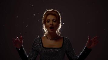 Beautiful singer in black dress performs on abstract, dark background in studio, with light shining down on her and snowflakes falling around her. video