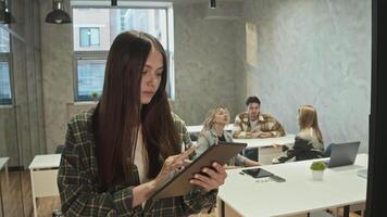 An attractive young student is seen using tablet as she studies in college. She is smiling and looking directly into camera, standing in classroom with students behind her. video
