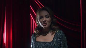 Beautiful and sexy pop singer, dressed in luxurious and shiny dress with precious stones, stands on stage in front of classic red theater curtain. audience in awe of her performance. video