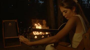 Young and attractive woman , in evening, sits by campfire with her friends. She adjusts strings of her out-of-tune acoustic guitar carefully, trying not to break them, she prepares to start singing with rest of group during their gathering. video
