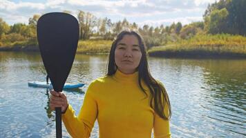 Portrait of young and beautiful Asian woman stands on shore, holding oar and looking at camera. She is preparing to stand up paddleboarding video