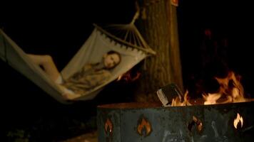 Young woman enjoys rocking and warming herself in hammock at night, lying next to brazier with bright flames from campfire escaping. Close-up video
