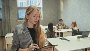 Portrait, young teacher with modern tablet in her hands looks at camera during break between lectures. happy university students are sitting behind it. video