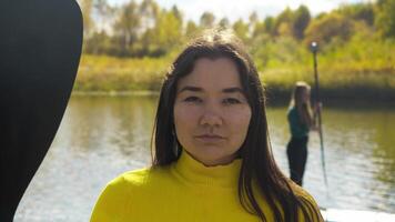 Pretty young woman, ready to go surfing, holds an oar in one hand and wears yellow sweater. She stands against backdrop of an autumn landscape with lake in distance. video