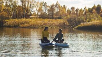 Two young girls are communicating with each other while doing my favorite activity - paddleboarding, in autumn landscape, floating on river. video
