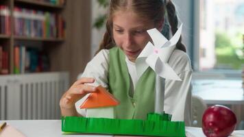 Happy smiling Caucasian little girl child school kid learner pupil student schoolgirl making handmade paper craft house home windmill ecology energy looking at camera showing thumb up indoor classroom video