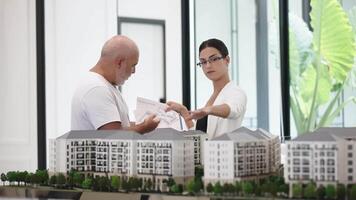 Business, architecture and office concept. Young people sharing plans and thoughts in office. Woman And Man Engineers With Model Of House Discuss About Design video