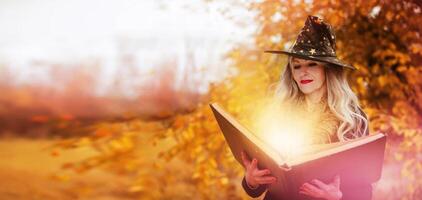 Halloween a woman with a magic spell book in a witch hat is conjuring, performing witchcraft over the autumn background of the forest. photo