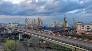 Overpass road and cargo train passing by railway under skyway. Maritime port landscape with yellow cargo cranes at waterfront. Slow motion pan view video