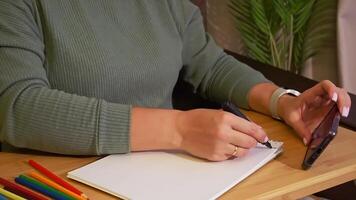Woman sketching on notebook while using smartphone at wooden desk video