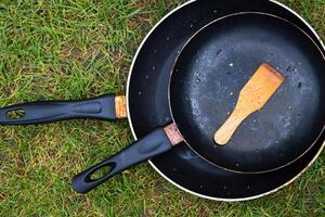 Top view of two black frying pans with a wooden spatula on a green grass, symbolizing camping lifestyle, outdoor cooking, minimalism, and rustic living concepts. photo