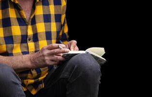 Closeup of a man in a yellow checkered shirt holding a pen and a cigarette, writing in notebook Smoking habits, creativity, note taking, journaling, planning concept photo
