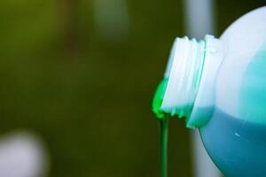 Closeup of vibrant green detergent pouring from a white plastic bottle against a blurred background. Green liquid flowing from a container. Copy space. Selective focus photo
