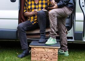 Casual moment of van life featuring two male friends. One man is smoking and writing in his journal, and another is browsing his smartphone, representing outdoor living, freedom, and minimalist travel photo