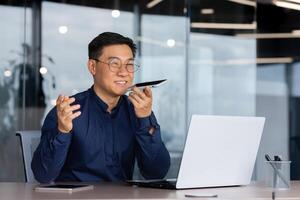 Successful asian man inside office working with laptop, businessman recording audio message using app on smartphone, man using artificial intelligence assistant to help finding a solution. photo