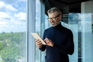 Mature adult businessman standing near window inside office, man smiling and using tablet computer, gray-haired boss browsing internet pages and reading news. photo