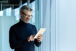 Mature adult businessman standing near window inside office, man smiling and using tablet computer, gray-haired boss browsing internet pages and reading news. photo